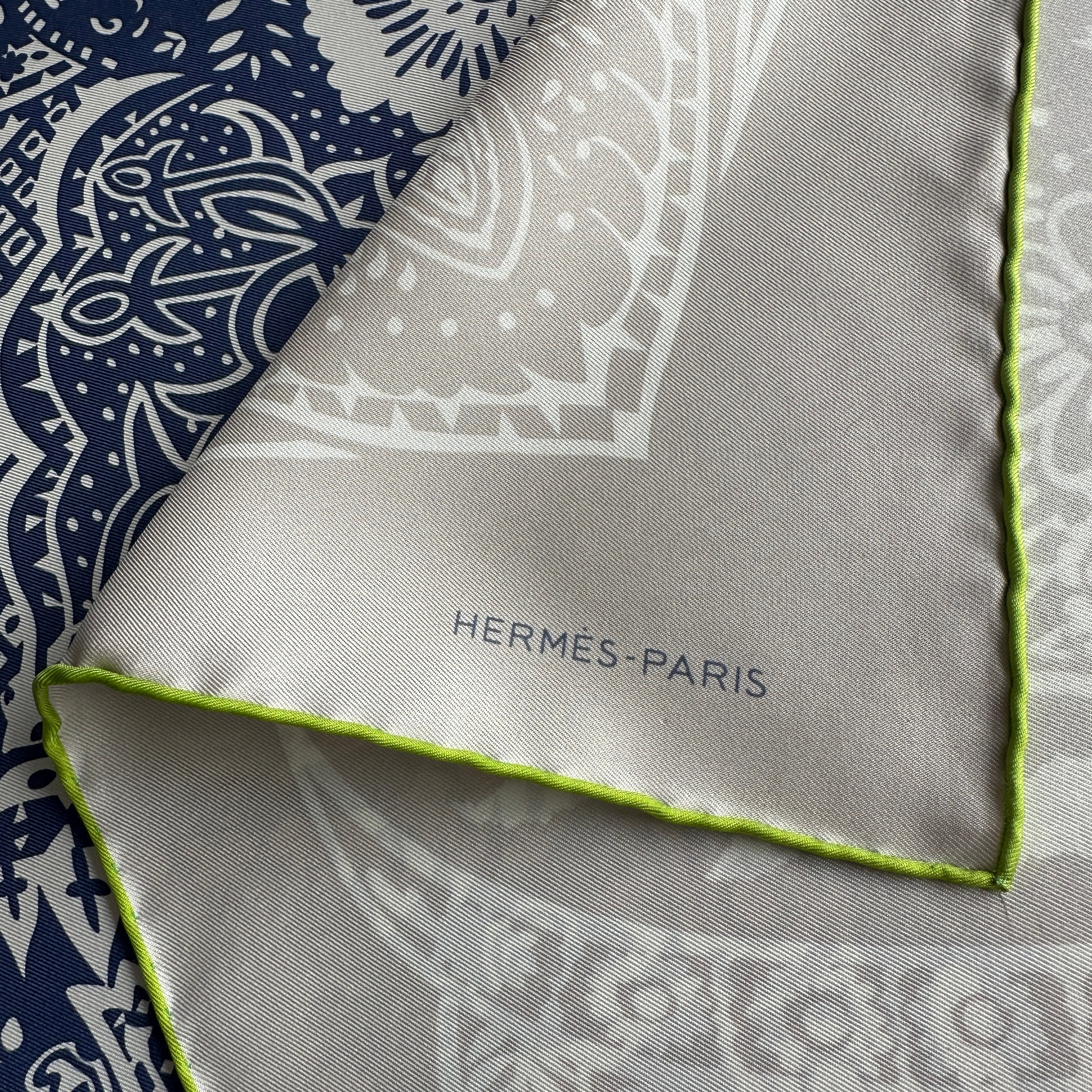 Zoom sur le coin du carré Hermès montrant la signature "HERMÈS-PARIS" imprimée sur la partie gris clair, près du bord roulotté vert lime.