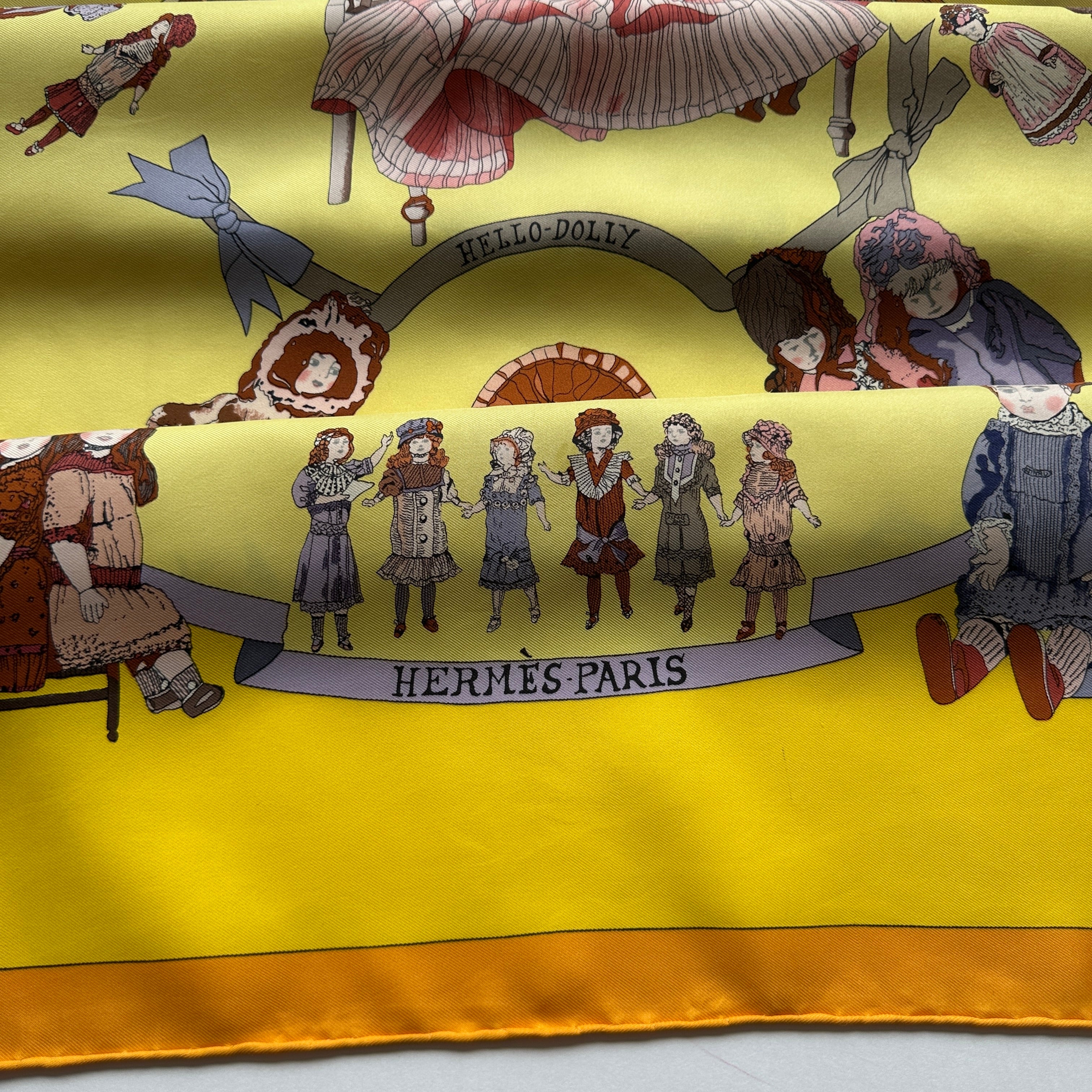 Inscription "HELLO DOLLY" sur un ruban gris et détail de l'ourlet roulotté main jaune orangé.